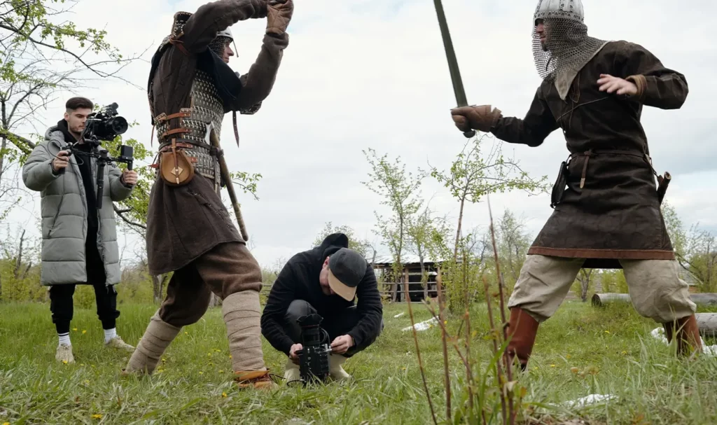 Two videographers film two medieval reenactors sword fighting in a field.