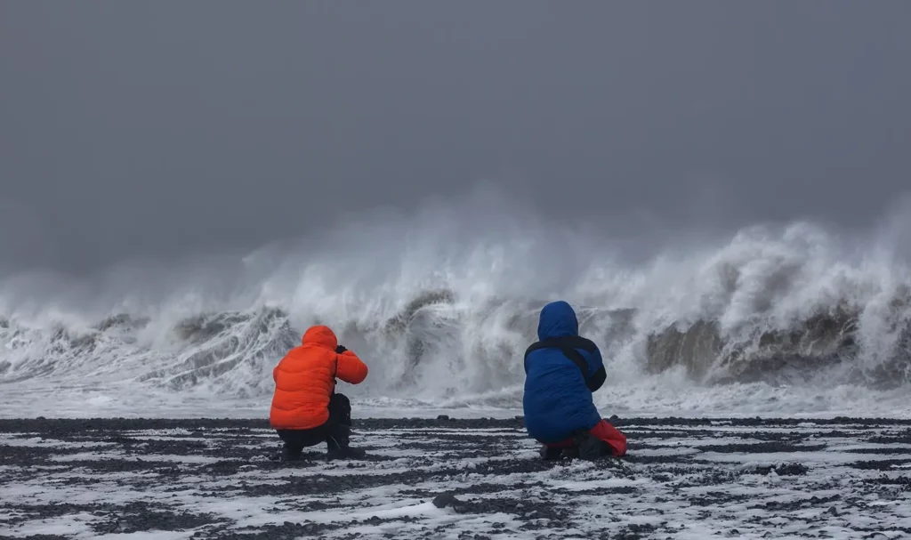 Two photographers in parkas film stormy waves on an Icelandic beach.