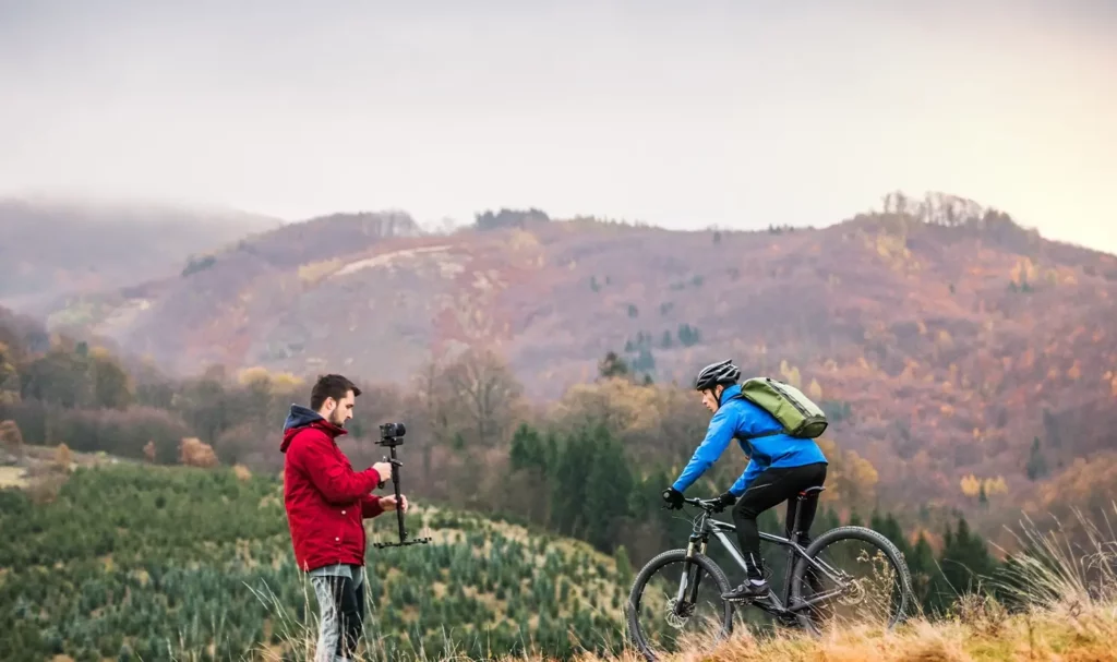 Videographer in a red jacket filming a mountain cyclist wearing a blue jacket and a backpack.