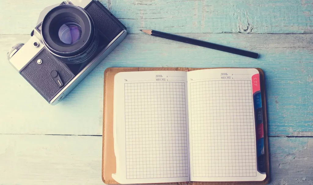 A vintage-style black and silver camera sits lens-up on a faded blue-gray wooden table next to a black pencil and an open planner with graph paper pages.