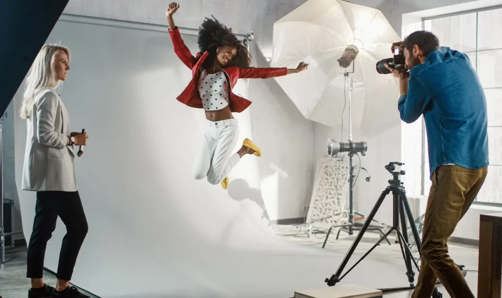 A photographer wearing a blue denim shirt and brown pants photographs a model, wearing a white outfit and a red jacket, jumping into the air in front of a white background. A makeup artist with blonde hair, wearing a white jacket, stands to the side.