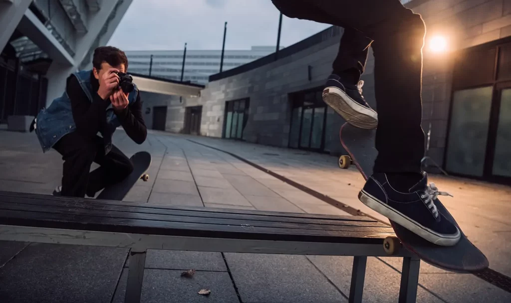 A photographer wearing black pants, a black long-sleeve shirt, and a denim vest crouches down while photographing a skateboarder wearing dark pants doing tricks on their skateboard.