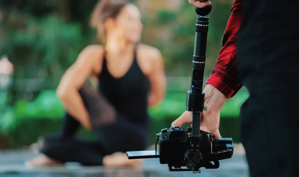 A close-up of a videographer's hands holding a camera on a gimbal, while filming a blurred yoga instructor in the background.