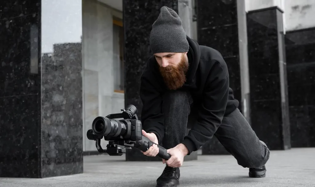 A bearded videographer wearing a dark gray beanie and all black clothes crouching near the ground while holding a video camera on a gimble.