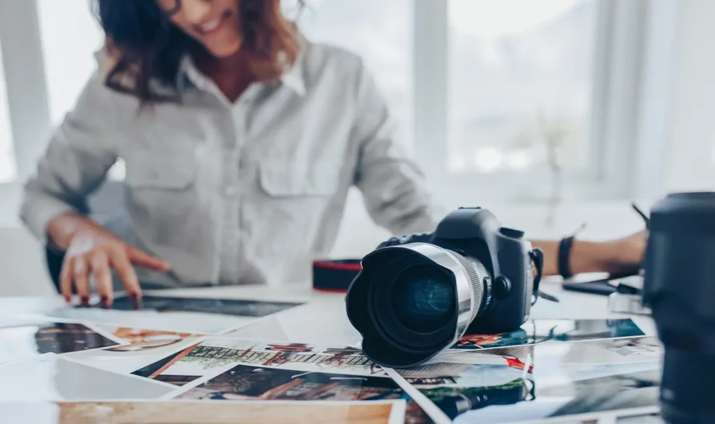 Tabletop covered in glossy photos with a camera in the foreground and a photographer reviewing photos in the background.