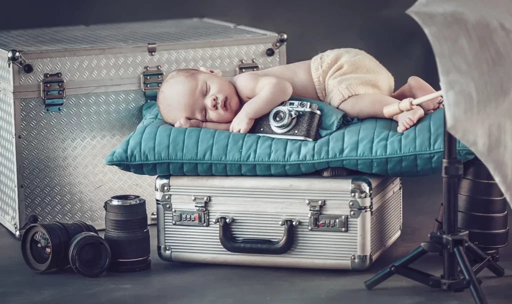 Newborn photo with baby laying on a teal pillow on top of a steel photography equipment case, with additional camera equipment around.