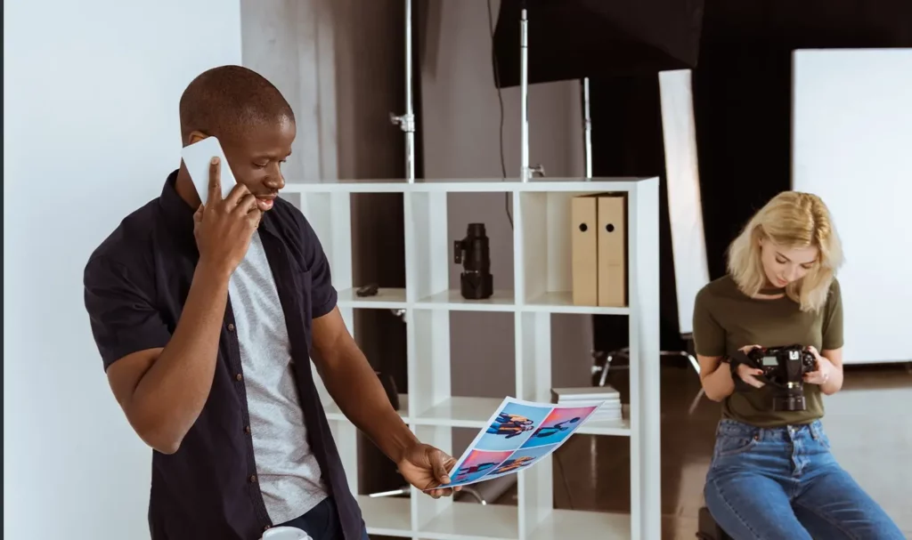 Male photographer wearing a dark, short-sleeve button-up over a white tee, talking on a smart phone and looking at photos, with a blonde model looking at a camera in the background.