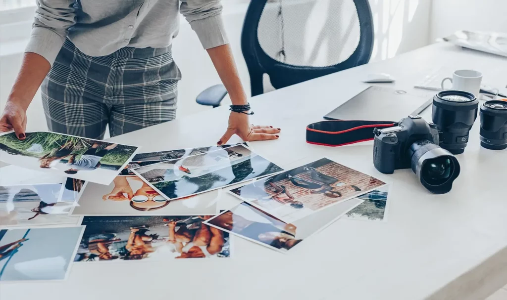 A white desk with a series of glossy photos spread across it, with several black camera pieces and a photographer's hands sorting through the photos.